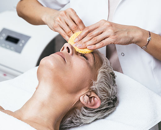 Beautiful senior woman having chemical peeling beauty treatment. The expert beautician is applying chemical solution onto woman's face.